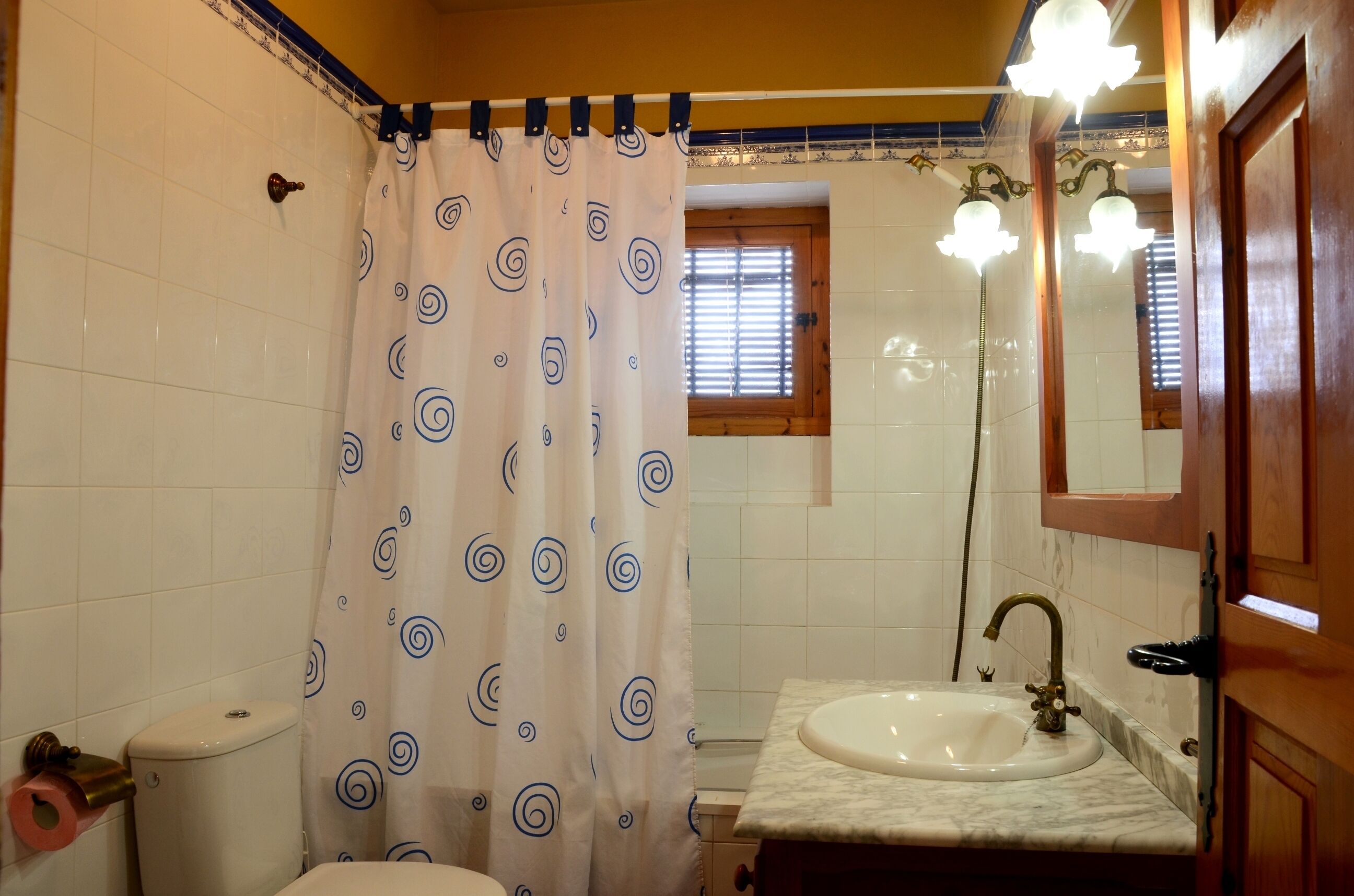 Et badeværelse med toilet, vask, bruser med gardin og et vindue med persienner.