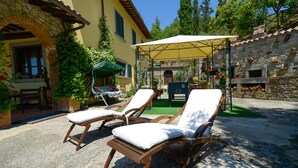 A courtyard with two wooden lounge chairs under a canopy, a stone wall, and a rustic outdoor kitchen area.