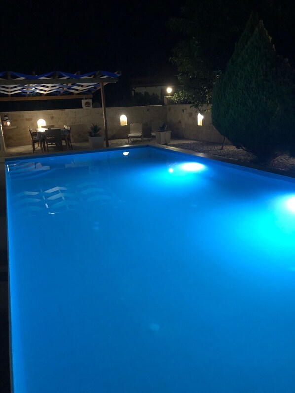 Une piscine dont le bord est illuminé de lumière bleue, entourée d’une terrasse avec une table et des chaises, et un mur agrémenté d’un éclairage décoratif.