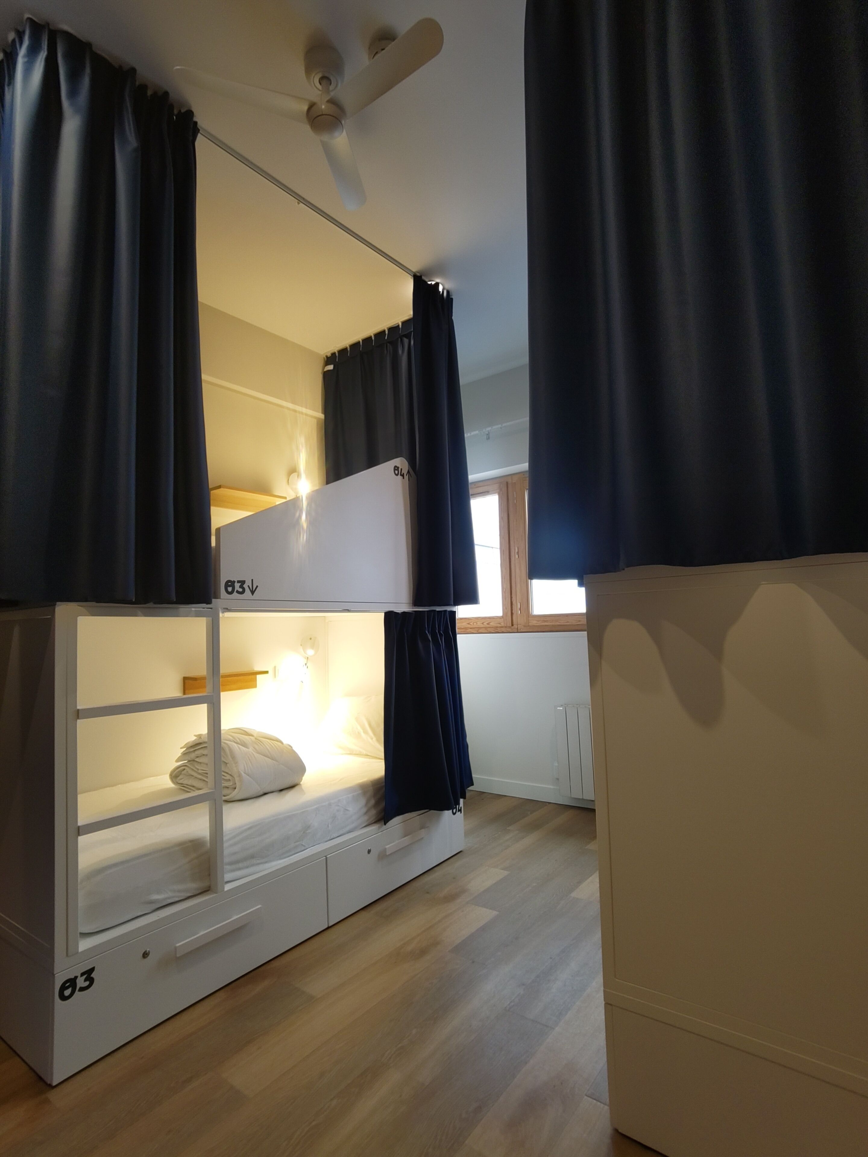 Una cama litera con cabecera blanca, ventilador de techo y ventana con cortinas.