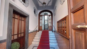 A grand hallway with a red carpet leading to a large arched doorway, flanked by wooden doors and a staircase with a red runner. - Lux comfort privacy in the center of Rome, shared by a group of 10 friends (Roma)