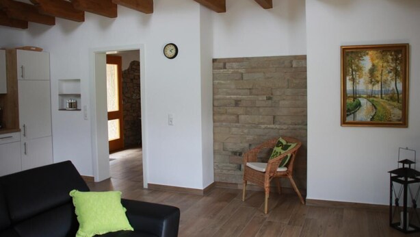 A living room with a black sofa, a wicker chair, a stone wall, and a painting of a landscape. - Enjoy the tranquility of nature, - / juniper reserve in Alendorf (Blankenheim /Alendorf)
