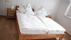 A wooden bed with white bedding and floral pillows, a wooden nightstand with a lamp, and a window with white curtains. - Enjoy the tranquility of nature, - / juniper reserve in Alendorf (Blankenheim /Alendorf)