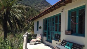A building with a white exterior and teal windows, a bench, and a palm tree in the foreground. - CASA AURA sunshine over the sea (Hermigua la gomera)