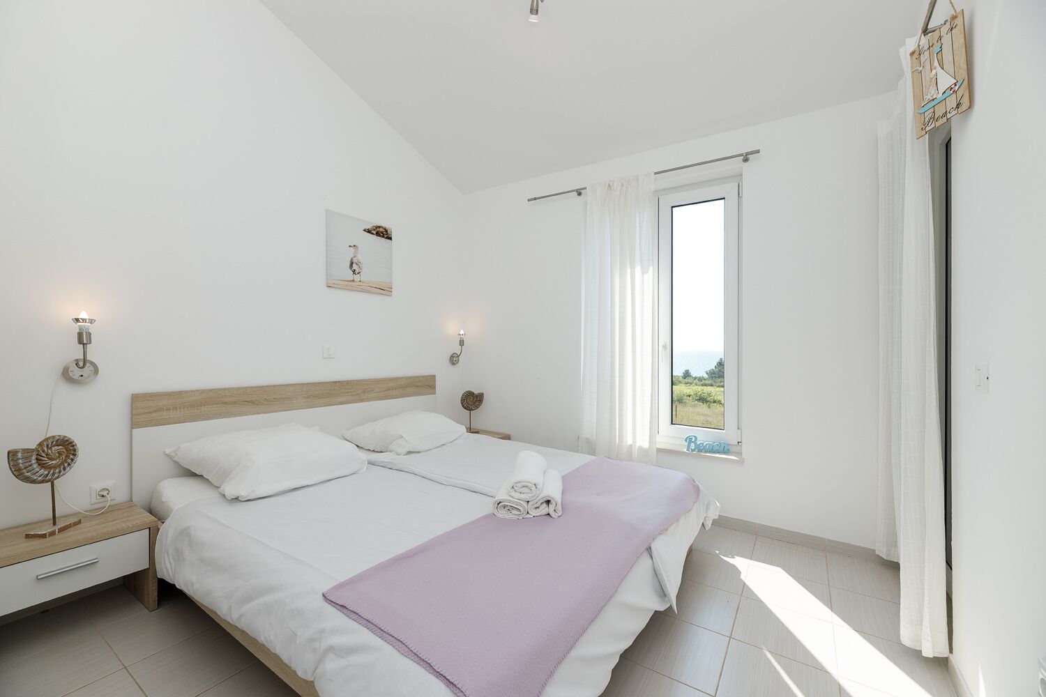 A bedroom with a bed, a nightstand, a wall-mounted light, a window with curtains, and a view of the sea.