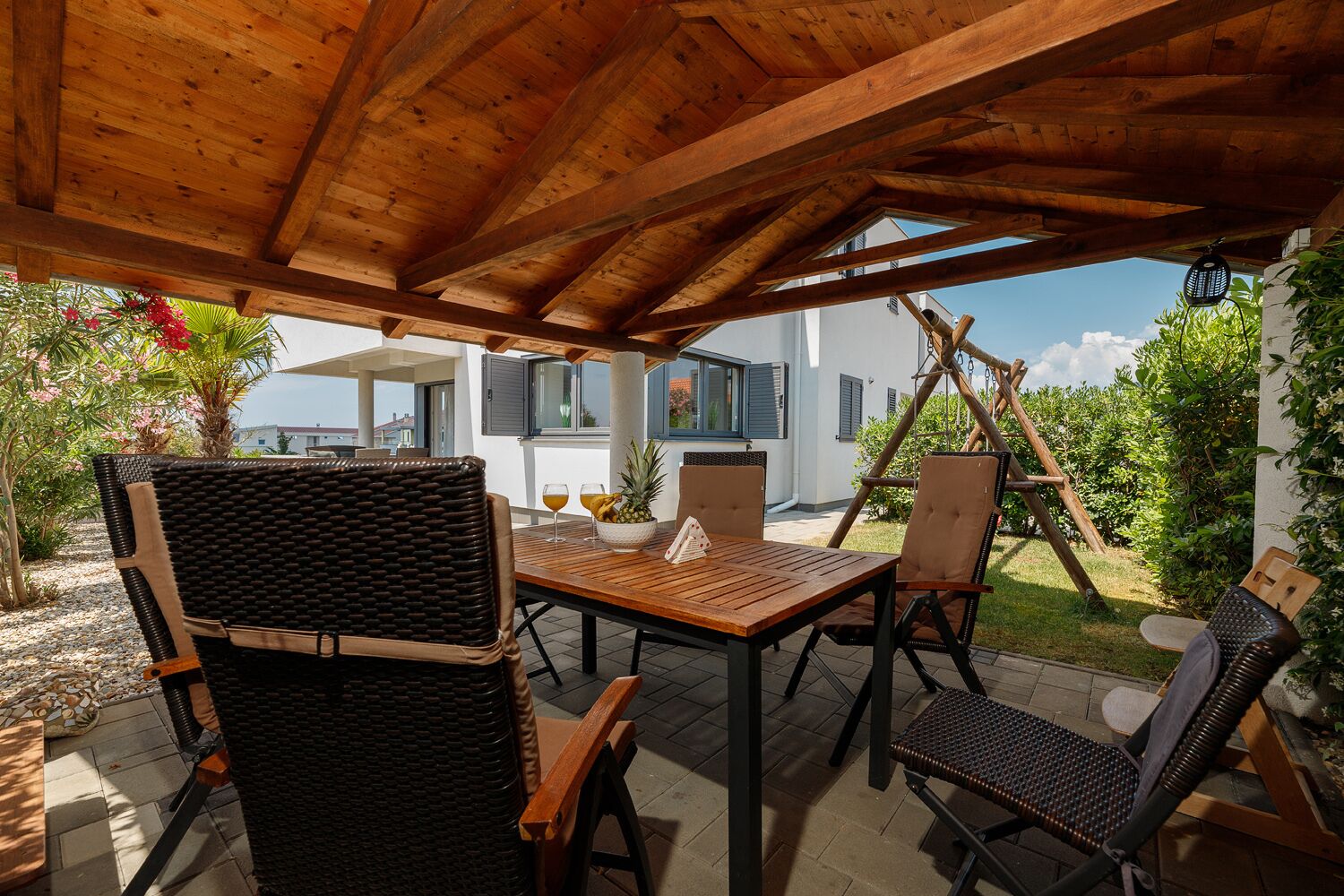 A covered outdoor dining area with a wooden table and wicker chairs, overlooking a residential building and lush greenery.