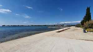 Une promenade en bord de mer, avec une eau d’un bleu limpide, un sentier pavé et une rangée d’arbres.