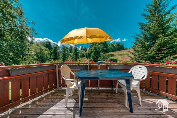 A wooden deck with a round table and two chairs, an umbrella, and a view of a forested hillside.