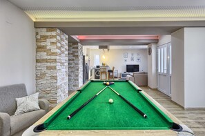 Ein Billardtisch in einem modernen Wohnzimmer mit einer Wand aus Stein, einem Sofa und einer Küche im Hintergrund.