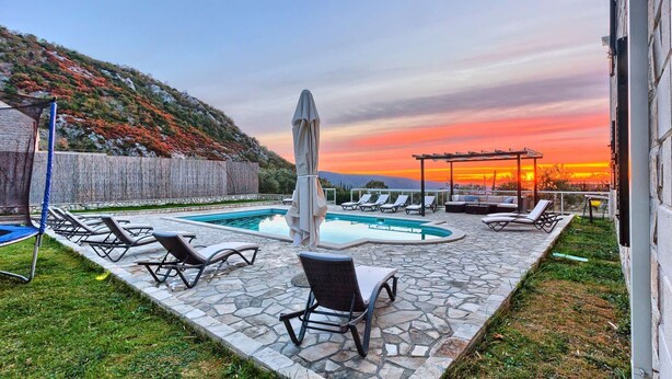 Un espace piscine doté de chaises longues et d’une pergola, entouré de carreaux de pierre et offrant une vue sur un coucher de soleil se déclinant sur un paysage vallonné.