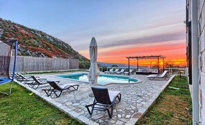 Ein Poolbereich mit Liegestühlen und einer Pergola, umgeben von Steinplatten und Blick auf einen Sonnenuntergang über einer hügeligen Landschaft.