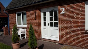 Un bâtiment en briques avec une porte blanche portant le numéro 2, un banc et deux plantes en pot.