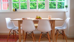 Uma área de refeições com mesa de madeira, cadeiras brancas e janela com vista para o verde.