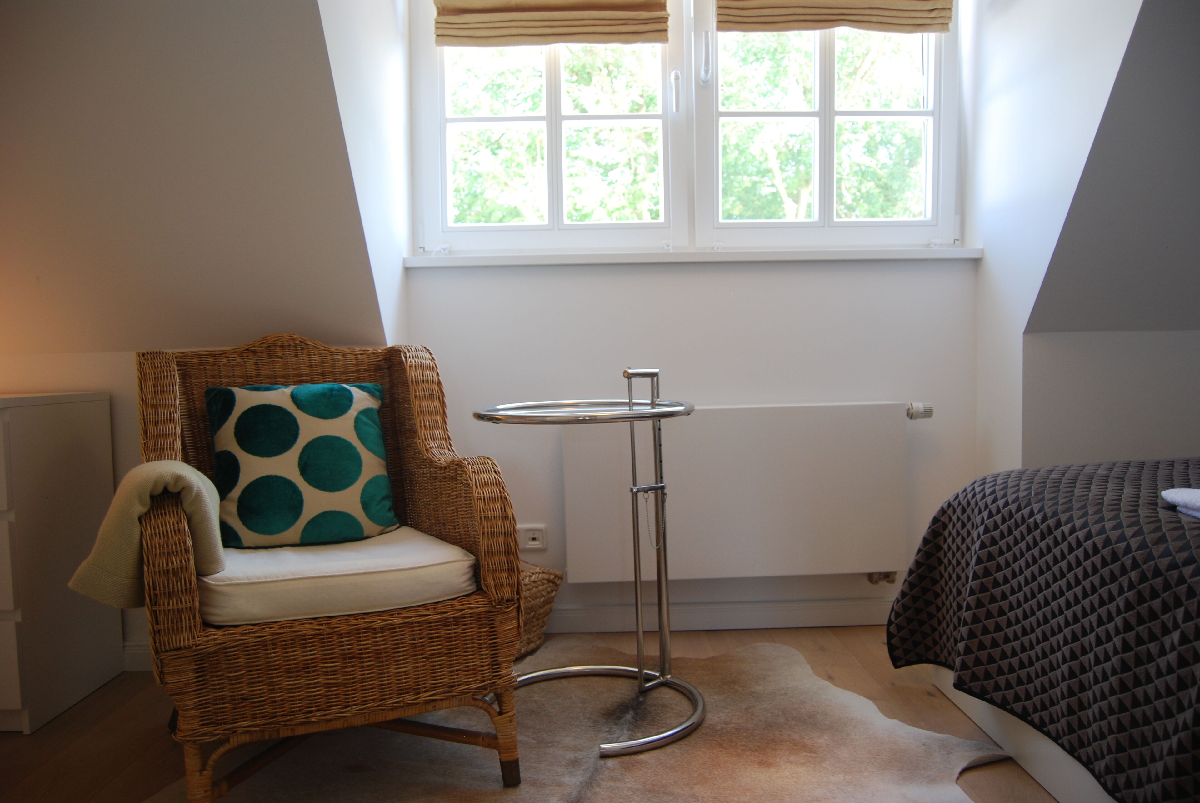 Een kamer met een rieten stoel met een blauwgroene kussen, een klein rond tafeltje, een radiator en een bed met een beddengoed met een geometrisch patroon.