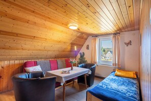 A cozy room with a wooden ceiling, a table with a vase, a sofa with cushions, a bed with a blue blanket, and a window with curtains.