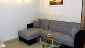 Ein Wohnzimmer mit einem grauen Sofa, einem Glastisch mit Blumenvase, einem gerahmten Bild an der Wand und einem schwarzen Lautsprecher.