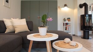 Ein modernes Wohnzimmer mit einem grauen Sofa, zwei weißen Beistelltischen, einer Topfpflanze und einem schwarzen Kaminofen.