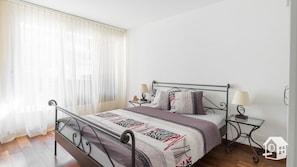 A bedroom with a metal bed frame, a large window with sheer curtains, a bedside table with a lamp, and a patterned bedspread.
