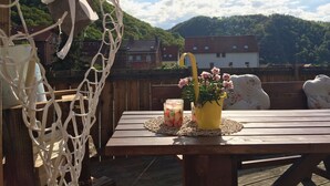 A wooden table with a yellow vase of flowers and a glass of juice on a wooden deck with a view of houses and hills in the background. - New Feriewohnung in the heart of the valley with stunning view to Roßtrappe, **** (Thale)