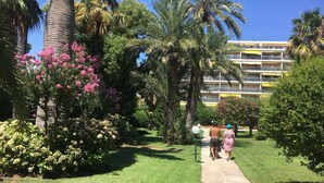 A pathway in a lush garden with palm trees and flowering bushes, leading to a multi-story building with balconies. - 4 *, BEACH / EVENTS, direct access to the beach, pool, garage, park. (Cannes)