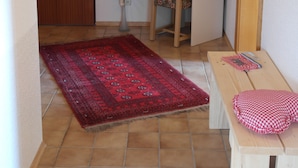 Una habitación con una puerta de madera, un escritorio con una impresora, una silla, una pequeña mesa con mantel a cuadros, una alfombra roja y un jarrón con flores.
