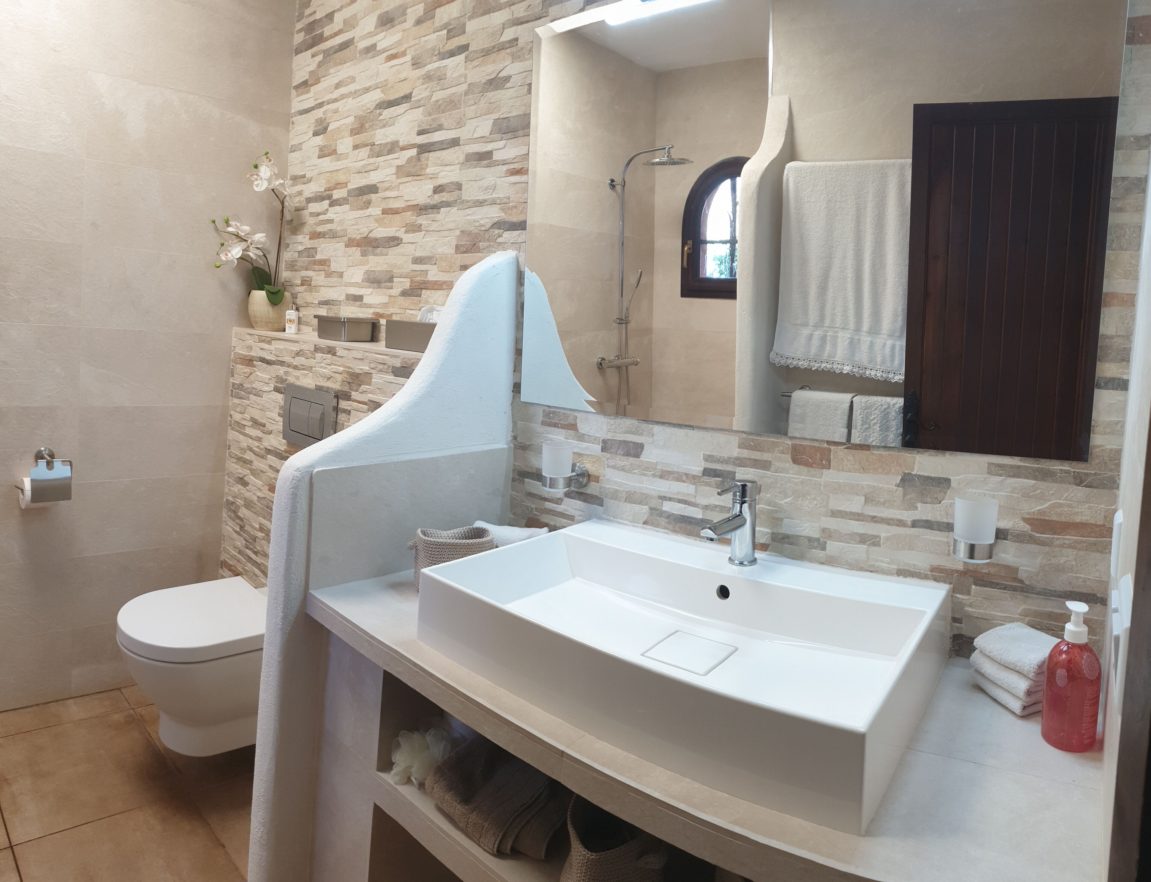 A bathroom with a white sink, a shower, a toilet, and a stone-patterned wall.