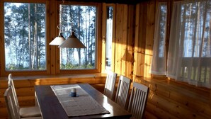 A wooden cabin with a dining table and chairs, large windows with curtains, and a hanging light fixture. - Cozy, 5 star log cabin directly on the lake. Water, forest, winter sports (Voipala)