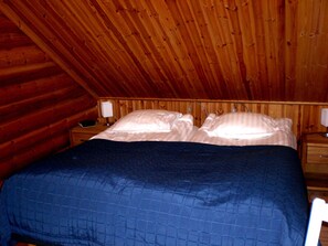 A bedroom with a wooden ceiling, a bed with a blue quilt, two bedside tables with lamps, and a wooden wall. - Cozy, 5 star log cabin directly on the lake. Water, forest, winter sports (Voipala)