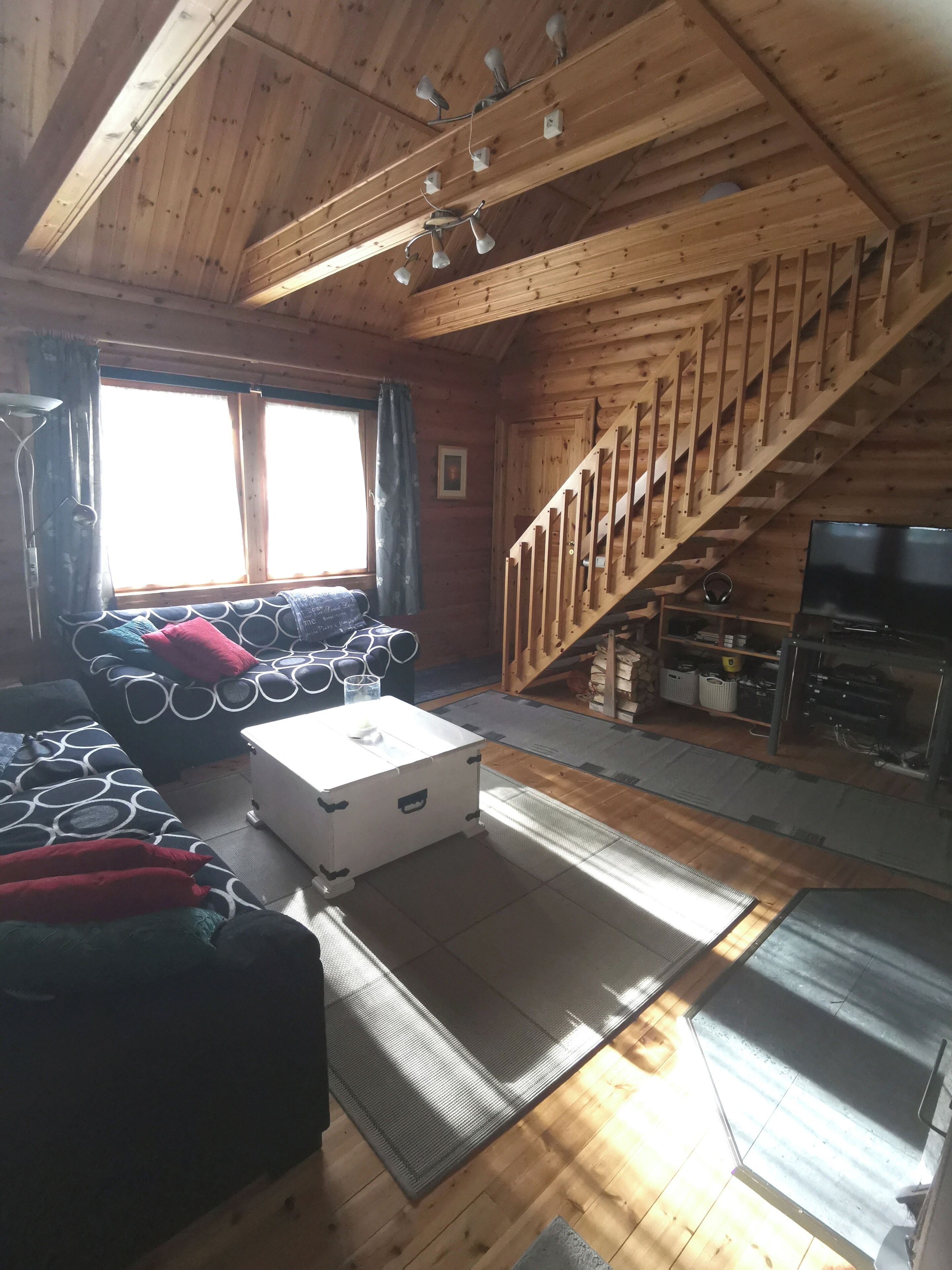 A wooden cabin interior with a loft bed, a white chest of drawers, a staircase, and a television.