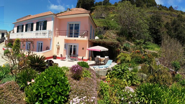 A two-story house with a balcony, surrounded by lush gardens and a hillside.