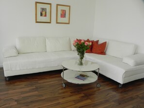 Ein weißes Sofa mit orangefarbenen Kissen, ein kleiner runder Tisch mit Blumen in einer Vase und zwei gerahmte Bilder an der Wand.