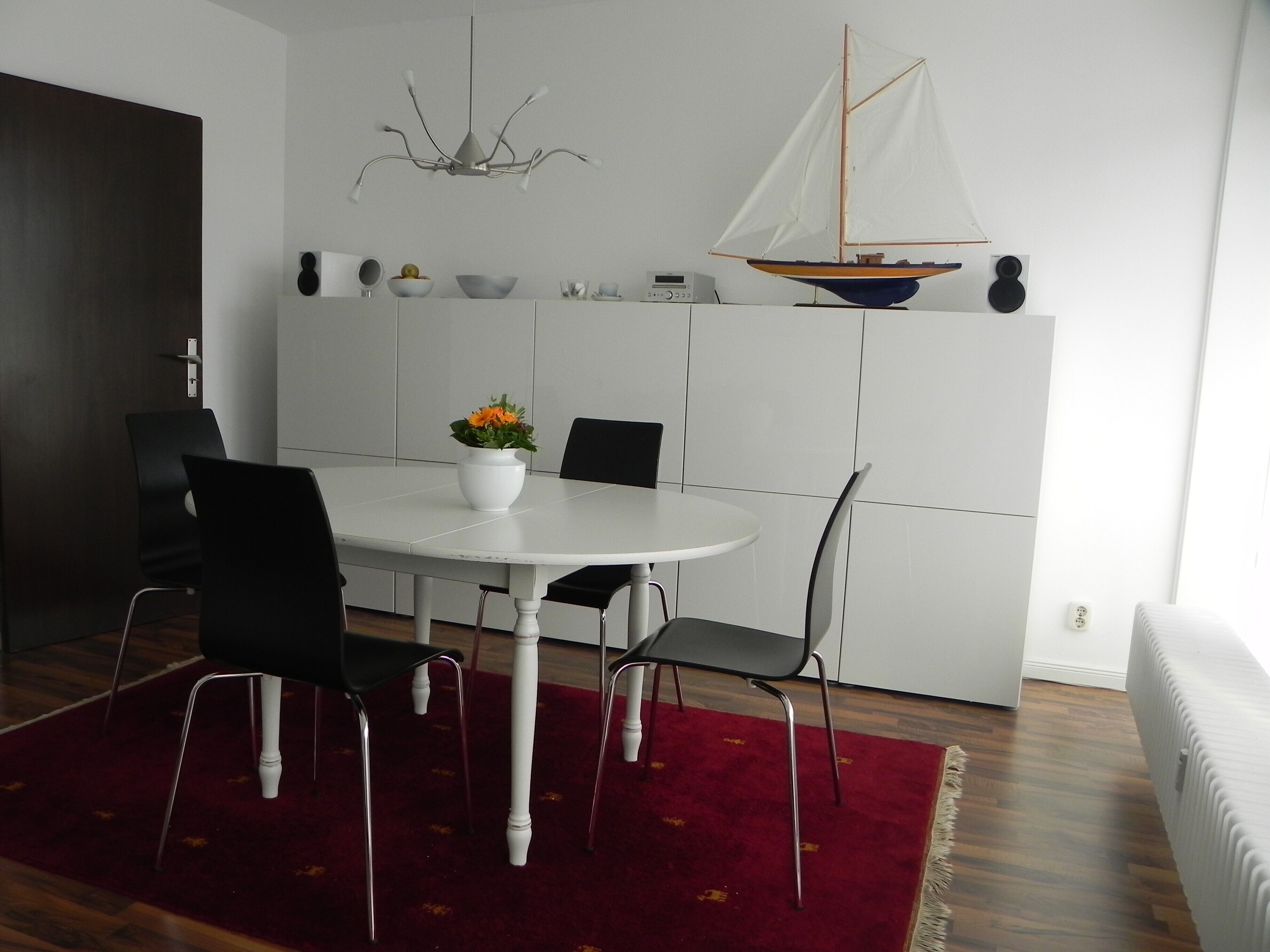 Et spiseområde med et rundt bord, sorte stole, en hvid vase med blomster, en sejlbådsdekoration og en rød måtte.