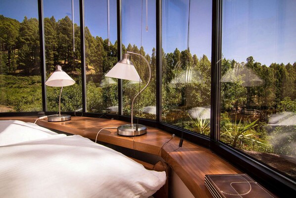 A bedroom with a large window overlooking a forested area, two bedside lamps, and a neatly made bed.