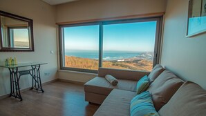 A room with a large window offering a view of the ocean and hills, a sofa with cushions, a glass table with bottles, and a mirror on the wall.
