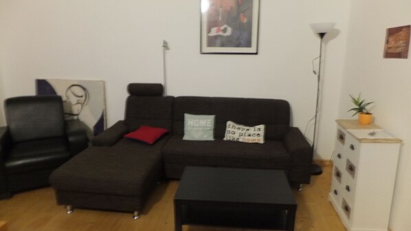 Ein Wohnzimmer mit einem dunkelbraunen Sofa, einem passenden Hocker, einem kleinen weißen Schrank und einer Stehlampe.