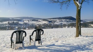 Dos sillas negras en un campo nevado con un árbol y una vista de un pueblo al fondo.