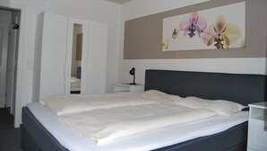 Una cama bien hecha con cabecera gris, una mesita de noche blanca con una lámpara y una puerta que da a un armario.