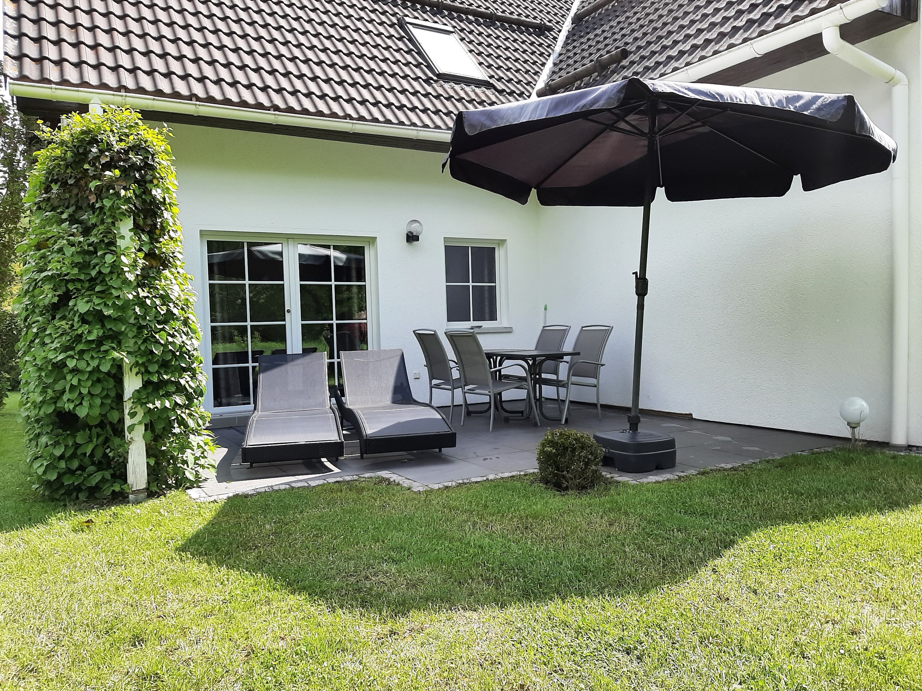 Une terrasse avec une table et des chaises, un parasol noir et un petit arbuste.