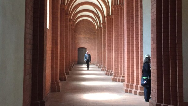 Un pasillo con columnas de ladrillo en arco y una persona caminando hacia una puerta.