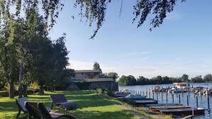 Un parque junto al lago con bancos, un muelle con barcos y un edificio al fondo.