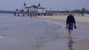 Een strand met een pier, een gebouw met twee torens en een persoon die langs de kust loopt.