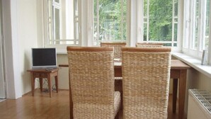 Een kamer met een houten tafel en stoelen, een laptop op een bijzettafeltje, en grote ramen met uitzicht op groen buiten.