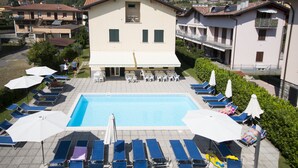 A pool area with lounge chairs and umbrellas, surrounded by buildings with balconies and a view of the surrounding landscape. - 3-room apartment to 6 persons with balcony  (Domaso)