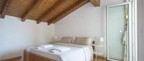 A bedroom with a wooden ceiling, a bed with a white bedspread, a bedside table with a lamp, and a sliding glass door to the right.