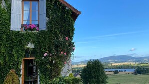 Ein Haus mit Holzhaufen und Fensterläden, dicht bewachsen mit Efeu und Blumen, mit Blick auf einen See und Hügel im Hintergrund.
