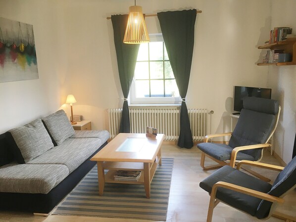 A living room with a sofa, a table, two armchairs, a lamp, a radiator, a window with curtains, and a television on a stand. - Apartment right on the dike (Butjadingen)