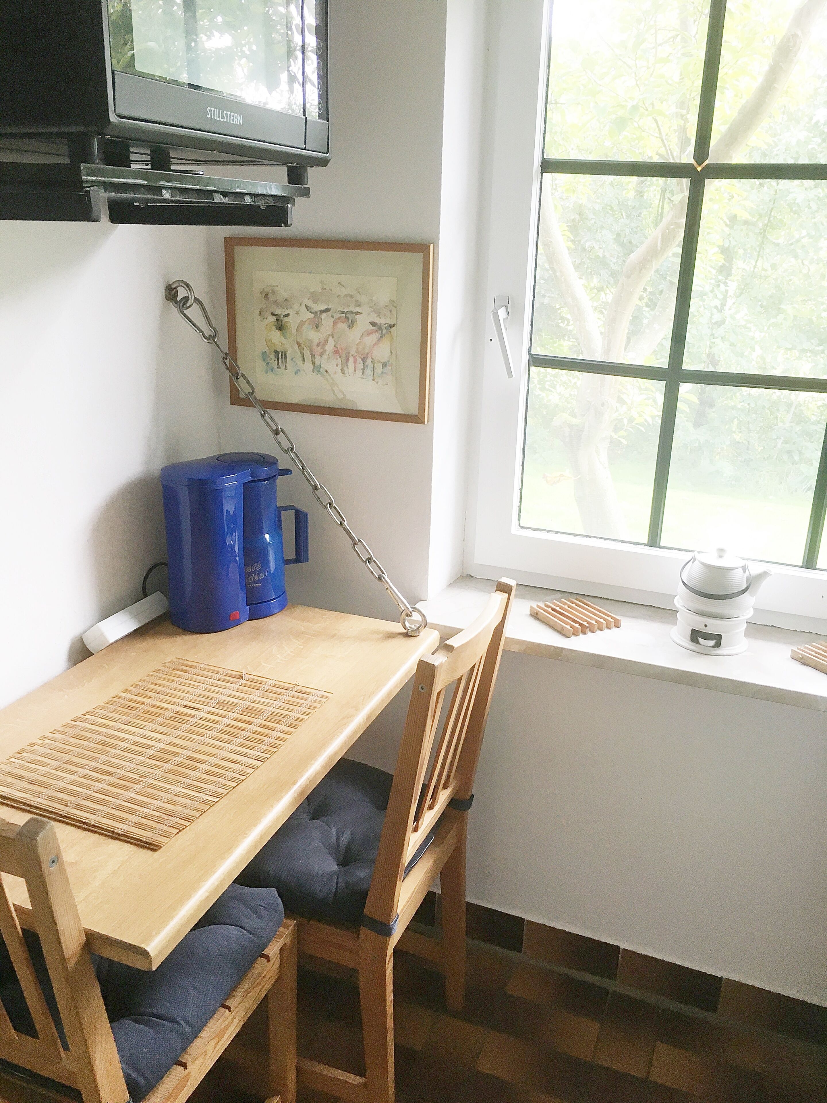 Een kamer met een houten tafel, stoelen, een televisie, een raam met uitzicht op bomen, en een schilderij in een lijst aan de muur.