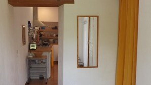 A hallway with a tiled floor, a wooden beam ceiling, a kitchen area in the background, and a yellow curtain on the right.