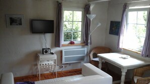 A room with a white sofa, a small white table, a wicker chair, a TV mounted on the wall, and a window with curtains.