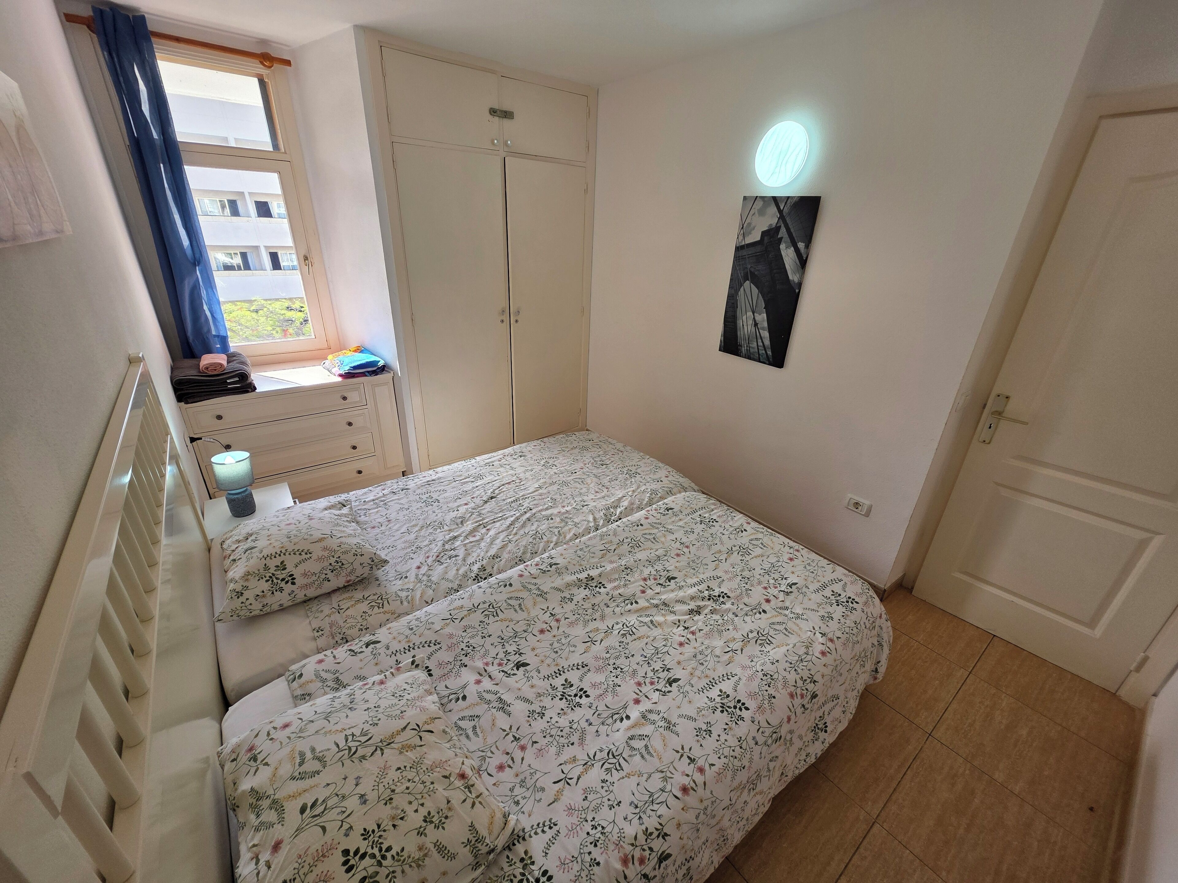 Ein Schlafzimmer mit Bett, Kommode, Kleiderschrank, einem Fenster mit Vorhängen und einer Wandlampe.